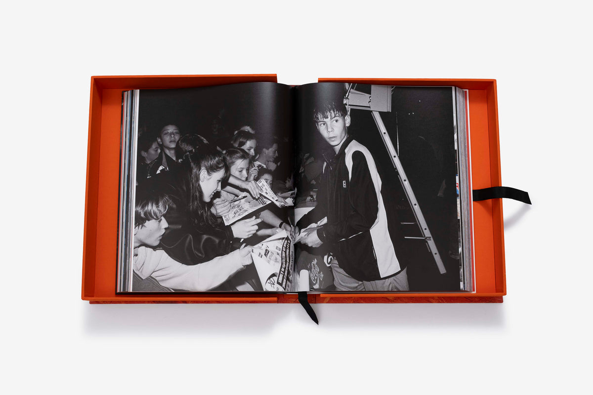 Rafa Nadal (Ultimate) by Assouline: the companion book open inside its orange presentation case to an archival black-and-white photograph of a young athlete in a track jacket signing autographs for a crowd of young fans