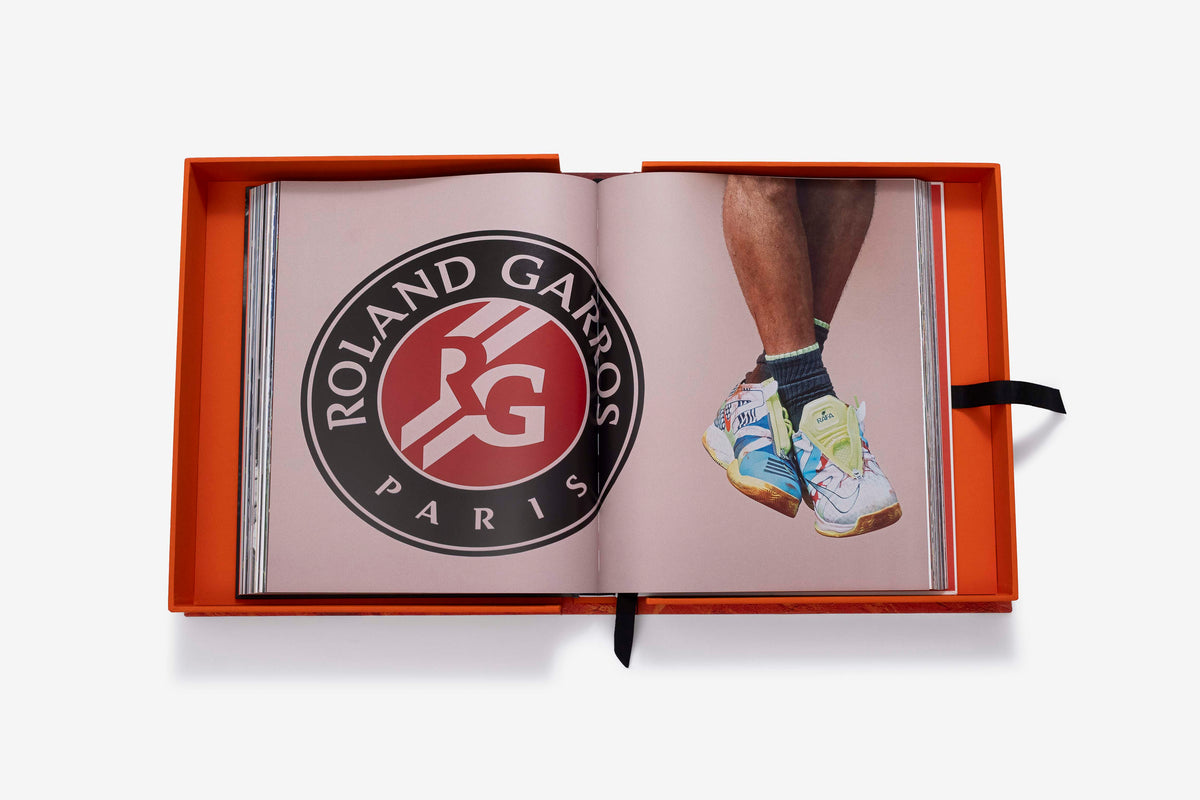 Rafa Nadal (Ultimate) by Assouline: the companion book open inside its orange presentation case to a pink-background spread showing the Roland Garros Paris logo emblem and a cropped photograph of a player's crossed legs and multicolor sneakers