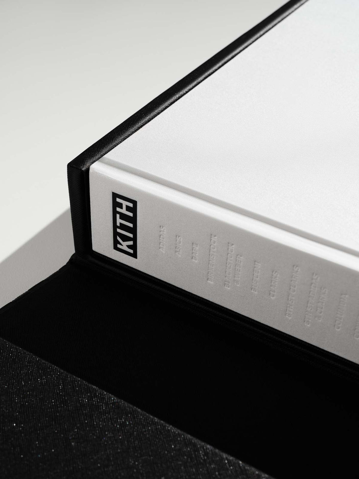 Close-up of a white Kith box with a black border and a black KITH logo on the side, resting on a black surface against a white background.