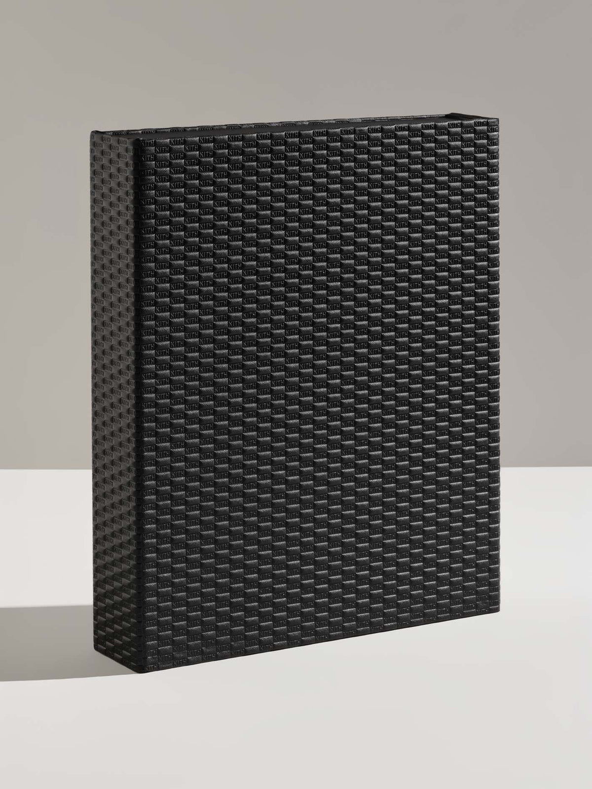 A black rectangular box with a woven wicker-style texture, standing upright on a light gray surface against a neutral gray background.