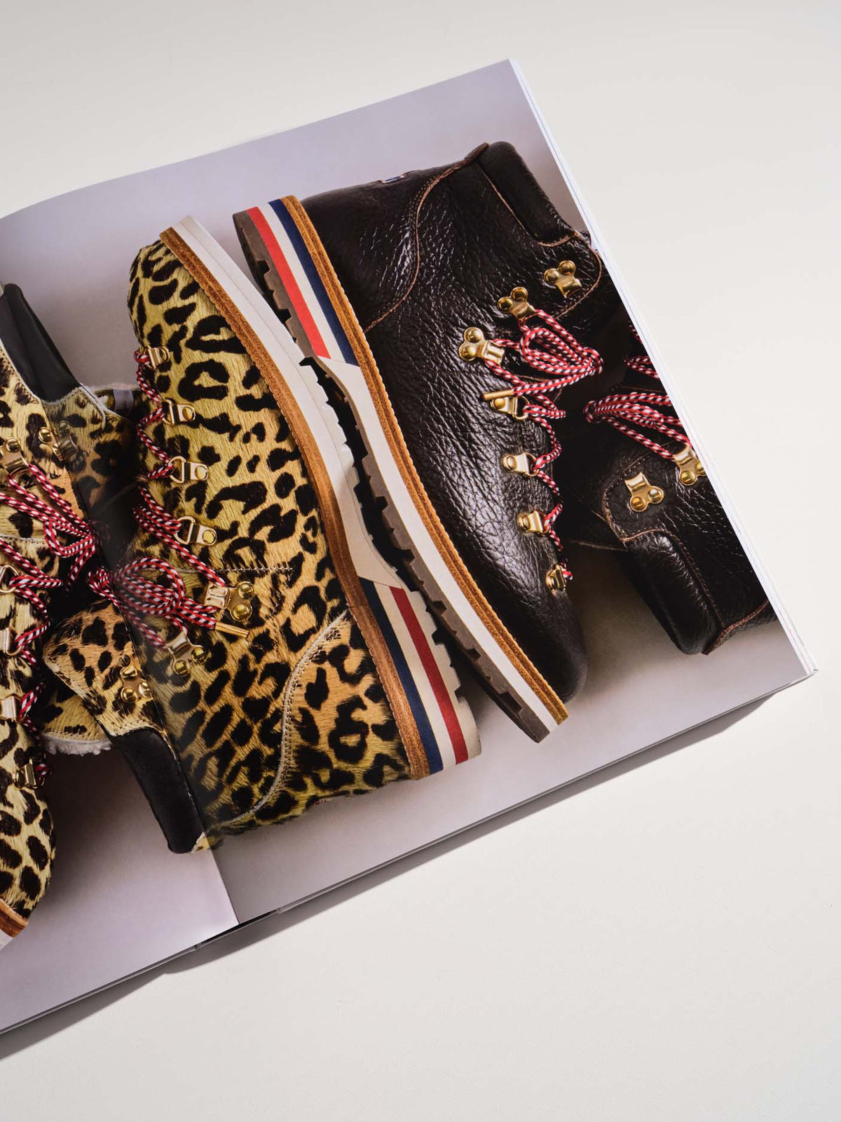 A pair of high-top boots displayed inside an open shoebox on a light gray background. The boots feature a dark brown pebbled leather upper with red, white, and blue striped soles and gold hardware, alongside leopard print boots with red patterned laces.