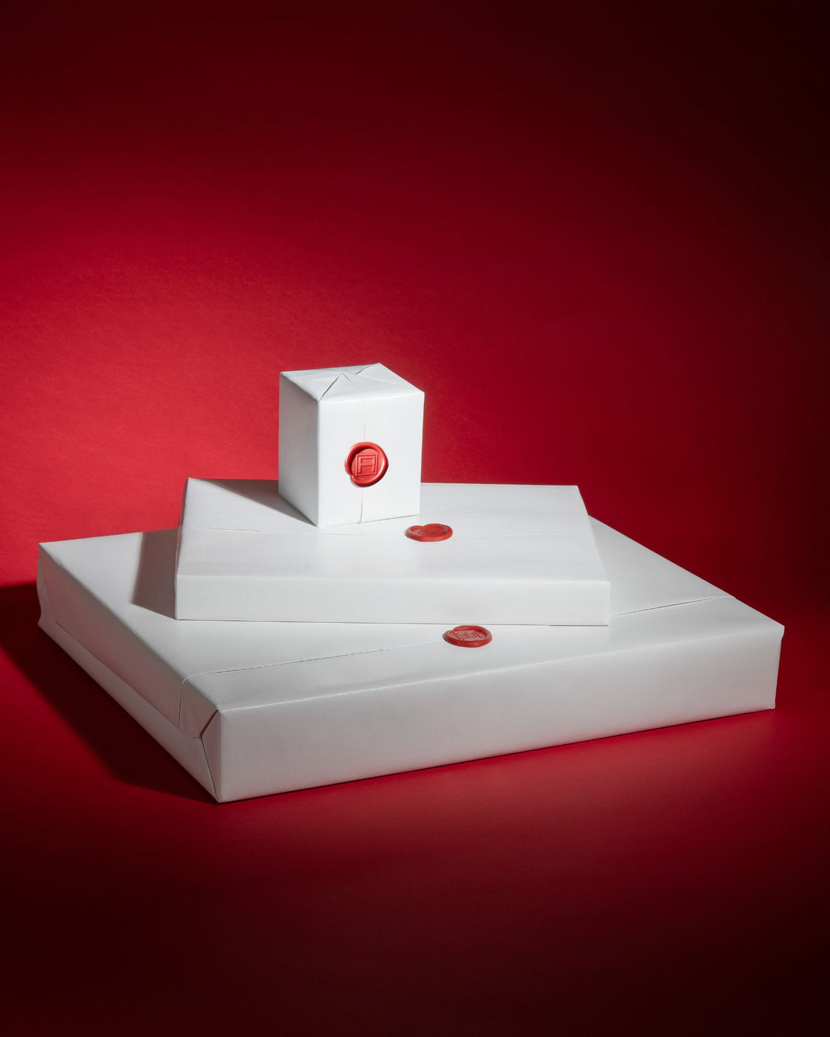 Three stacked white rectangular boxes with small red circular seals, photographed on a deep red studio background.